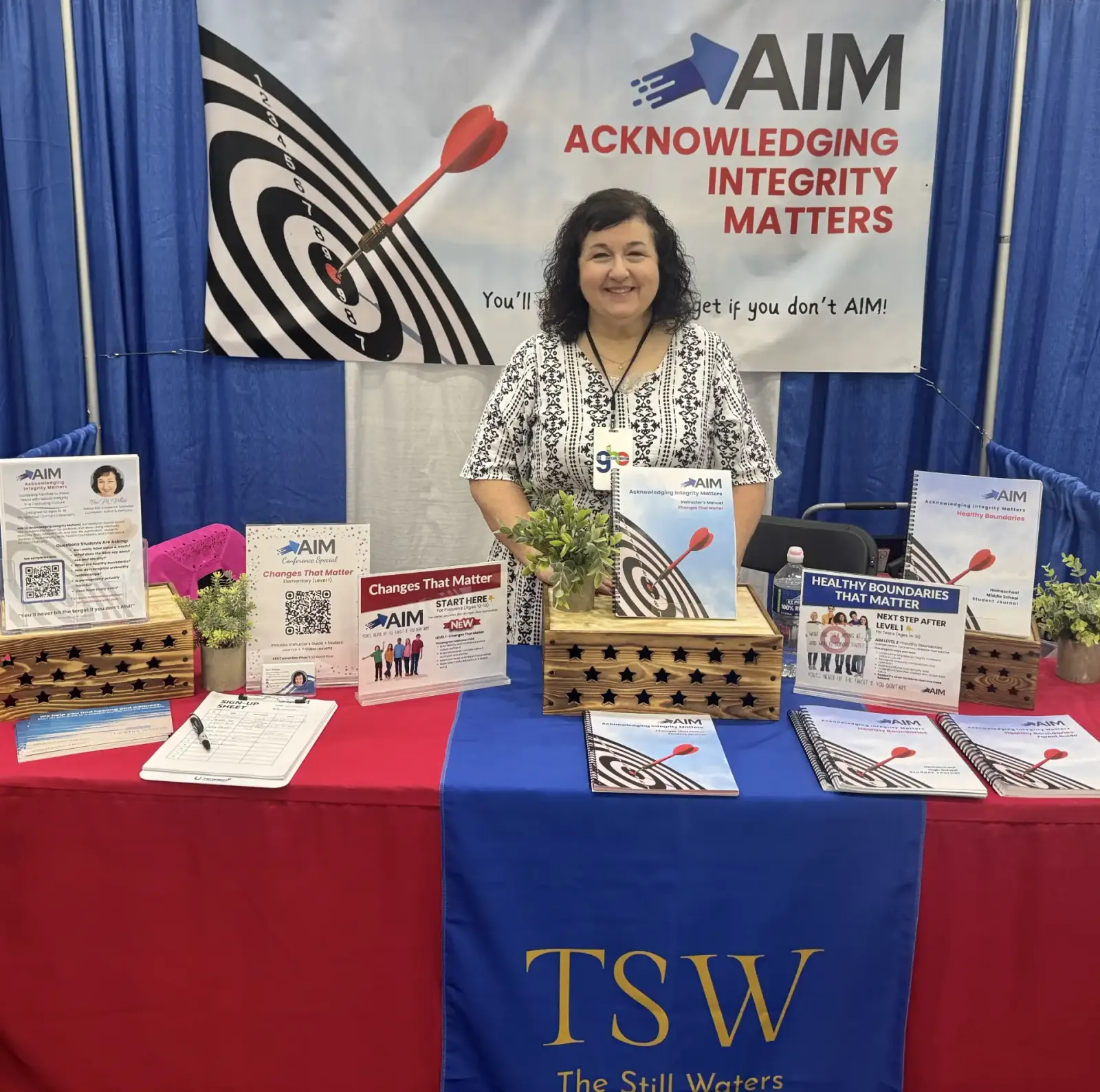 Teri White standing behind the AIM exhibit booth, with the AIM "Acknowledging Integrity Matters" target banner and curriculum materials on display