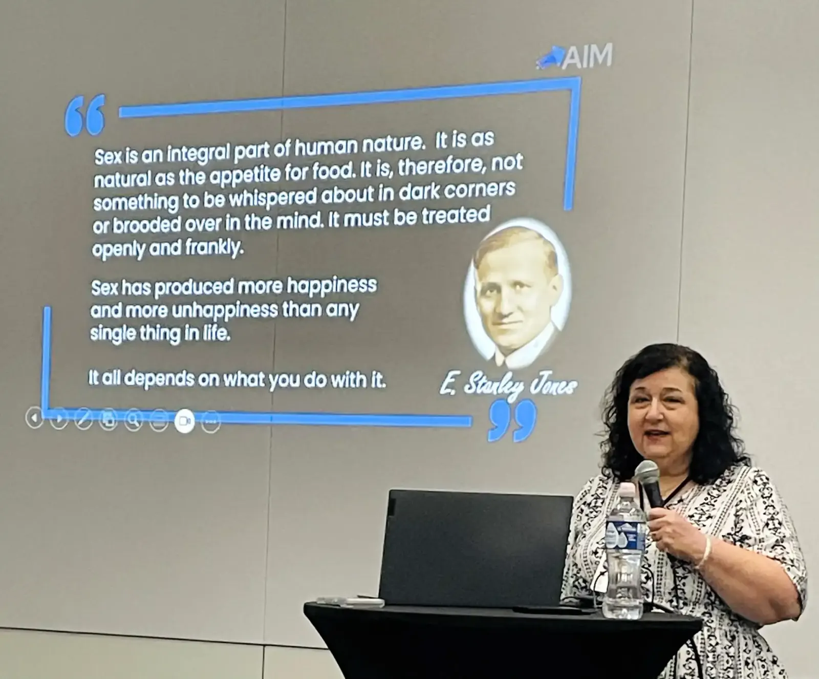 Teri White speaking at a conference podium, presenting an AIM slide with a quote on sexual integrity from E. Stanley Jones